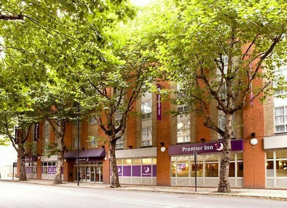 Outside of Premier Inn London Tower Bridge Hotel with purple signs, brick building with windows and grass, pavement and road in front