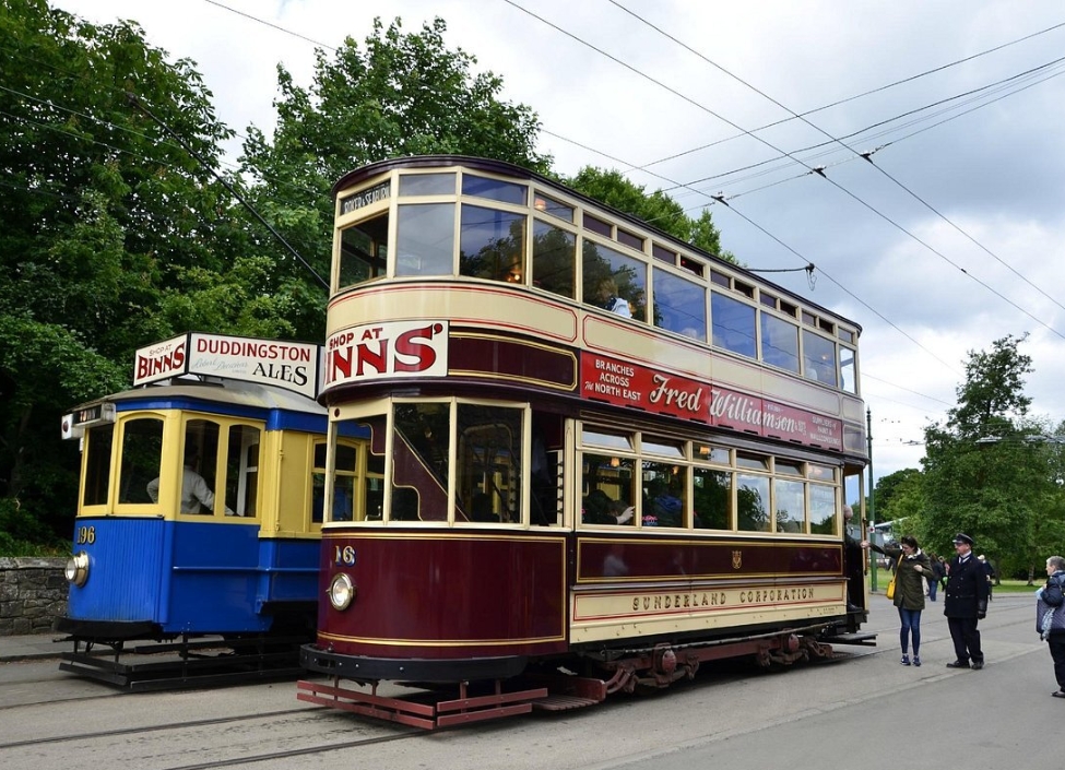 Two trams on Rails at Beamish Museum in Durham with oassengers and a conductor stood on the road.