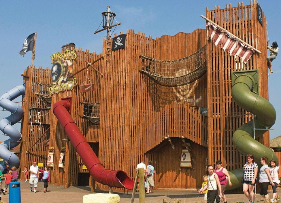 A wooden climbing frame at Crealy Theme Park withslides and rope bridges, with people walking around.