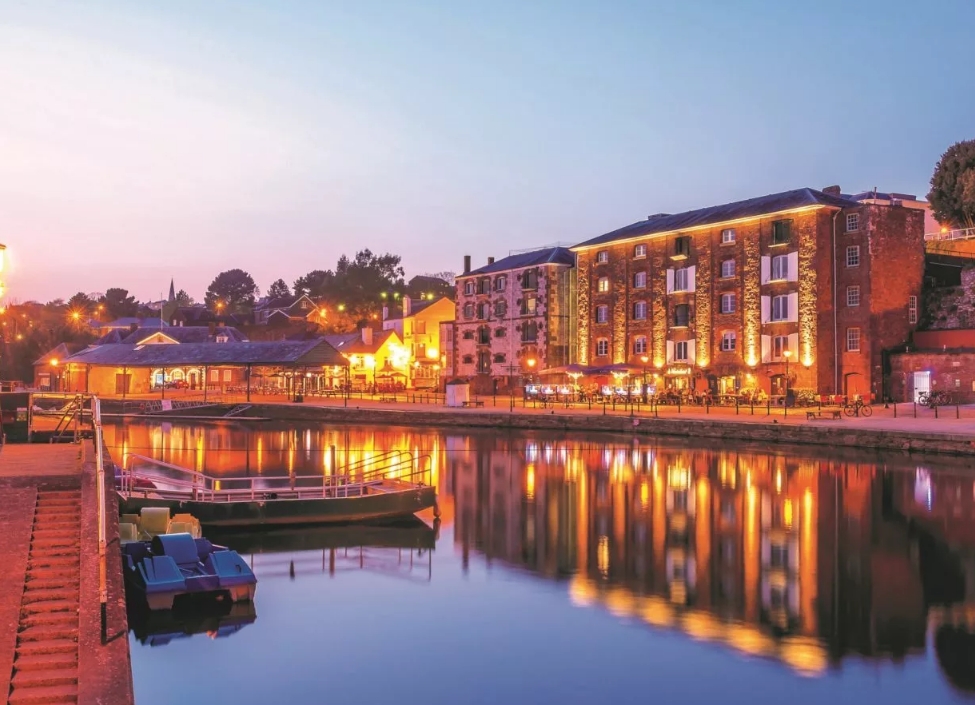 Exeter Quay at night with buildings on the side by a walkway and boats in the River Exe.