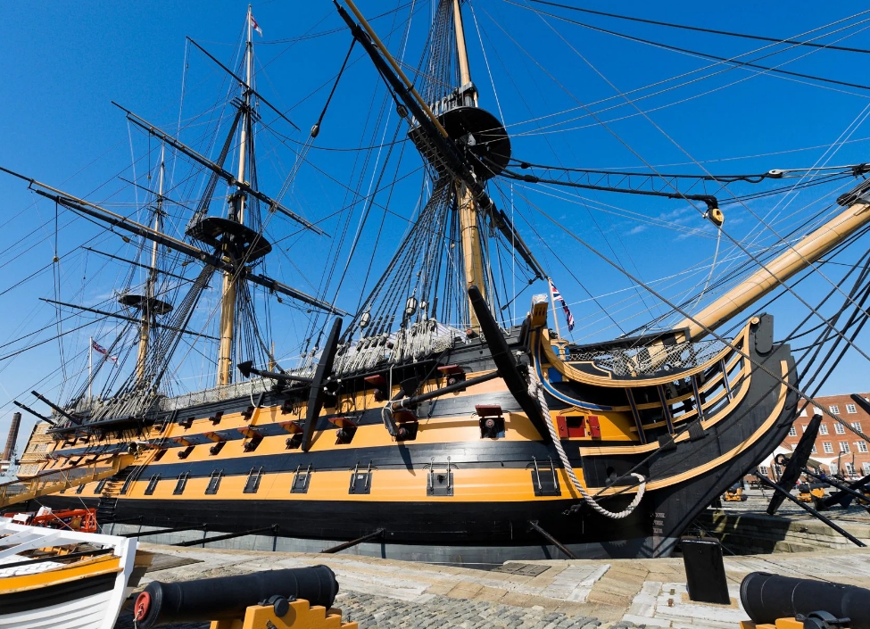 The HMS Victory, a yellow and black tall ship in Portsmouth Dockyard with pavement in front.