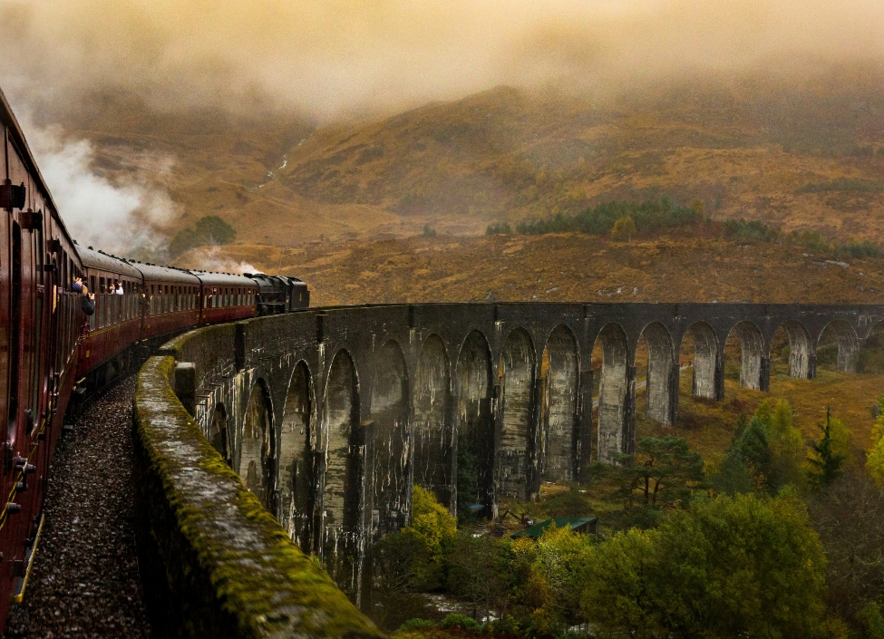 A red Jacobite Steam Train passing over the Glenfinnan Viaduct with trees and grass surrounding it
