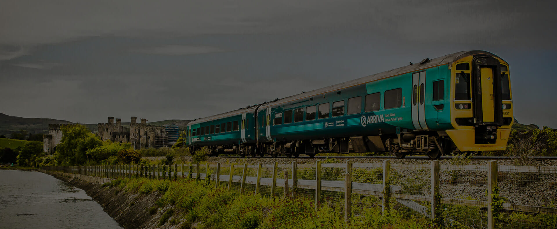 arriva trains wales green and yellow train travelling between stations in country side with castle in the background and fencing alongside