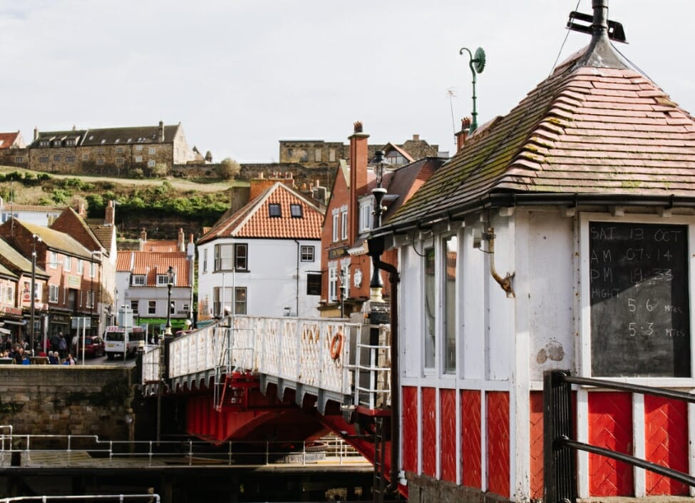 whitby harbour with shops and houses in the background and a red and white bridge in the foreground