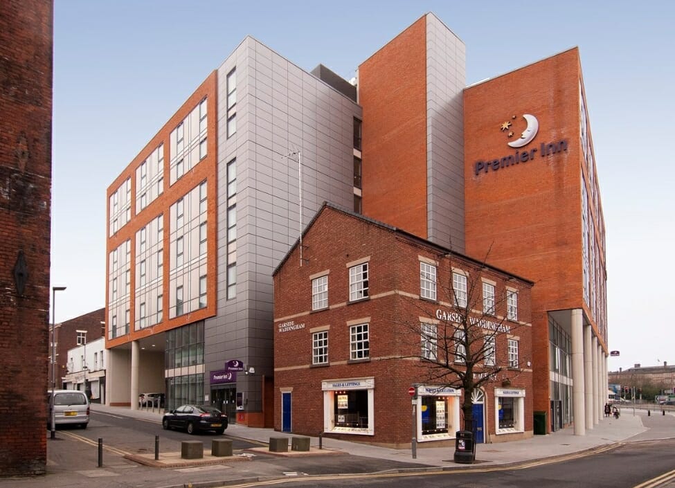 The Premier Inn Central Hotel in Preston, a red blick building with cladding with a smaller building surrounding it, and a pavement and road in front