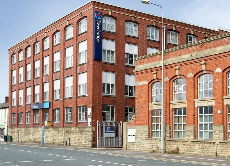 Travelodge Preston Central Hotel, a tall red brick building and a smaller building separated by a road with a pavement and road in front