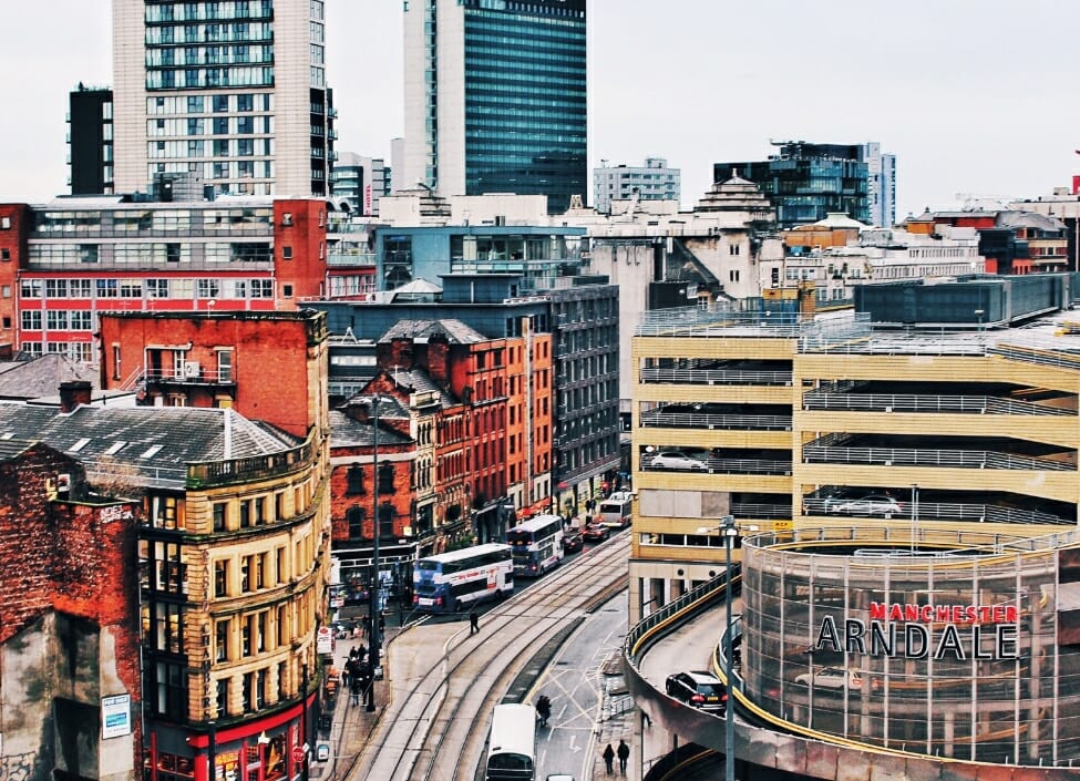 Manchester Arndale surrounded by other buildings with a road with buses and taxis and a tram line next to it