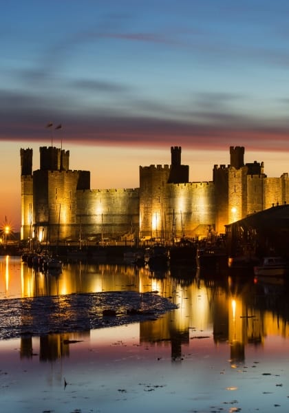 Caernarfon Castle in Wales lit up at night with water in front and an orange and blue sky behind.