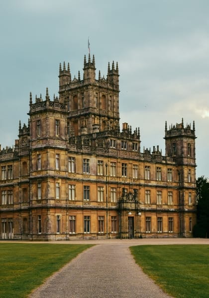 Highclere Castle, a tall yellow limestone building with numerous windows and turrets and pavement and grass in front.