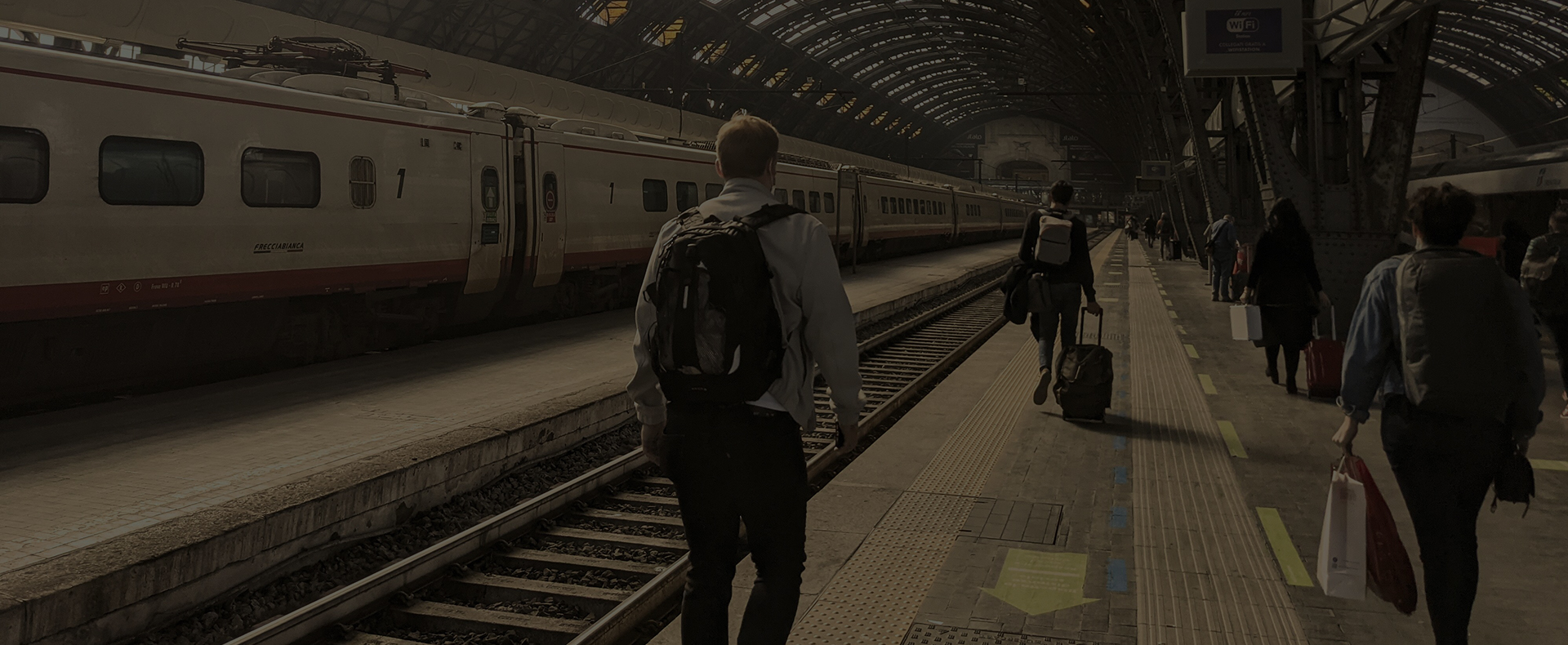 Passengers walking along a train platform with luggage, with a train waiting on the next platform.