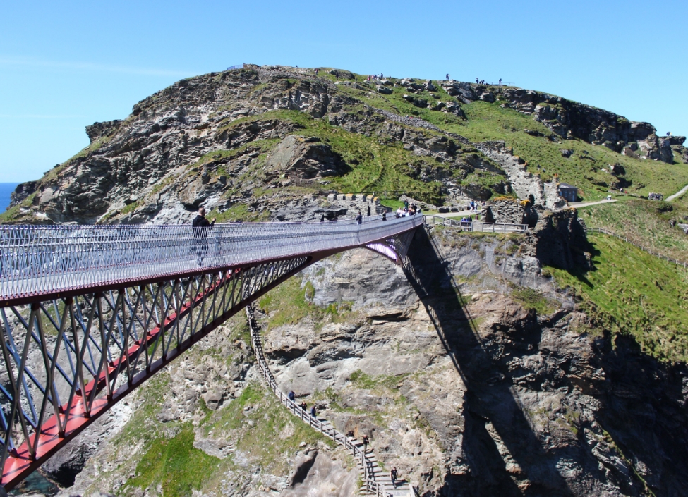A metal bridge crossing at Tintagel Castle with people walking across and rocky terrain with steps below