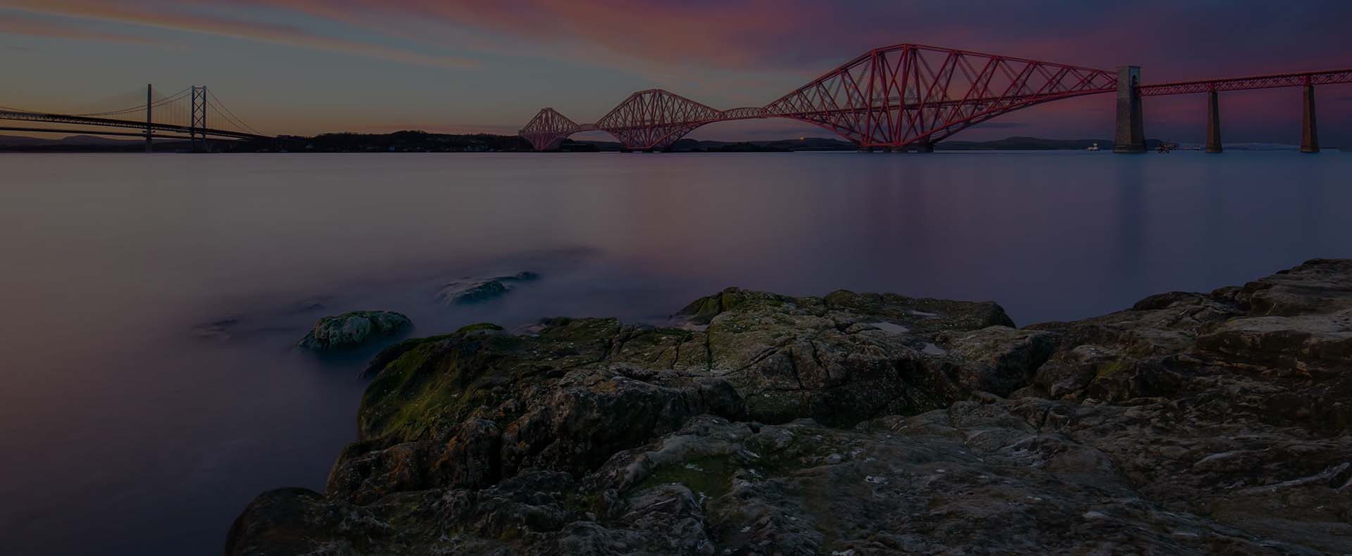 Forth bridge, a red railway bridge in Edinburgh with another bridge in the background and rocks and sea in the foreground