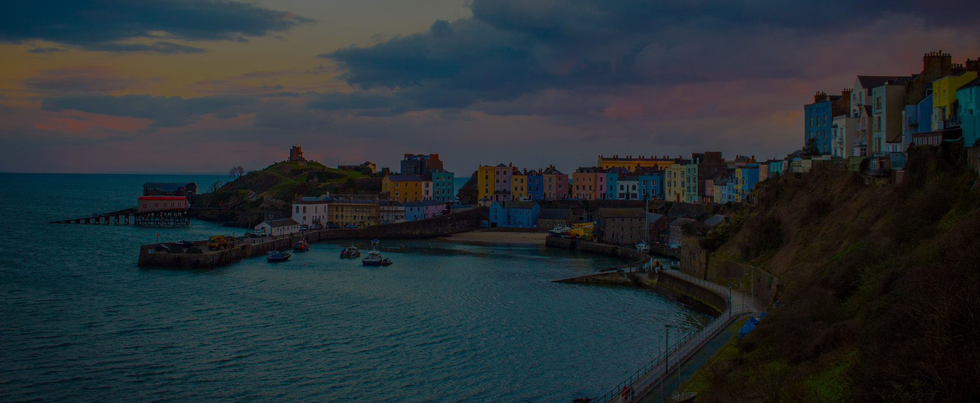 Tenby Harbour in Wales with colourful buildings lining the hill, a small beach, and a path running below grass.