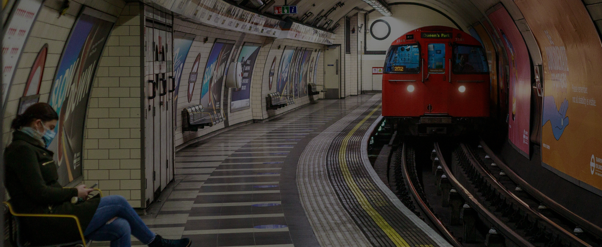 A tube to Queen's Park approaching a platform at London Waterloo Tube Station witha passenger sitting and waiting at the platform.
