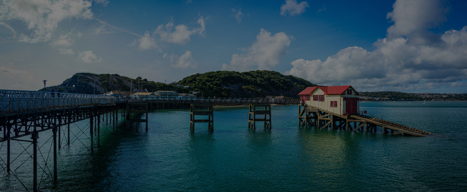 The Mumbles pier in Swansea over the sea with a red and white building at the end and a wooded area behind.