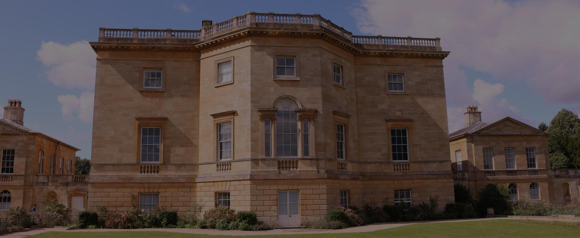 A sandstone manor building in Lower Basildon, Reading with multiple large windows and railings on the roof, surrounded by two other buildings and plants.