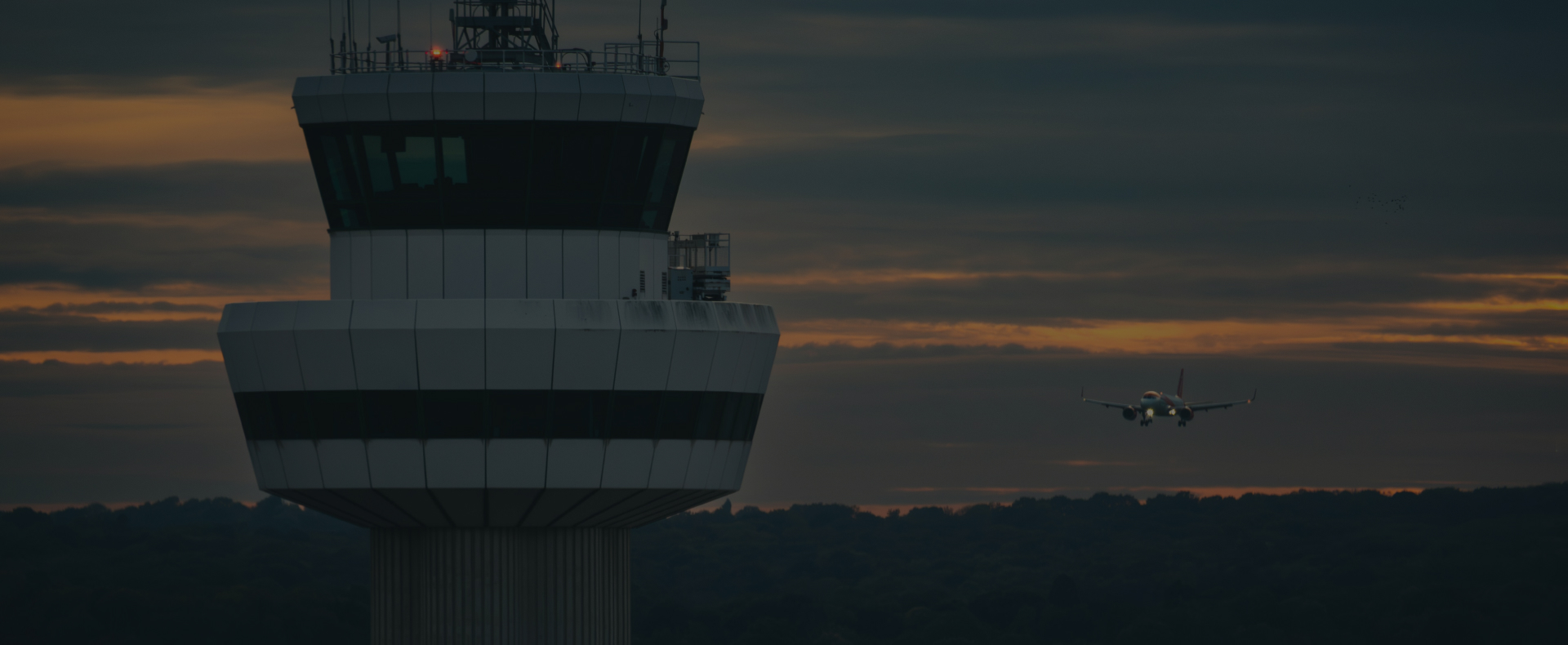 The control tower at Gatwick Airport with a plane approaching in the background and a cloudy sky.