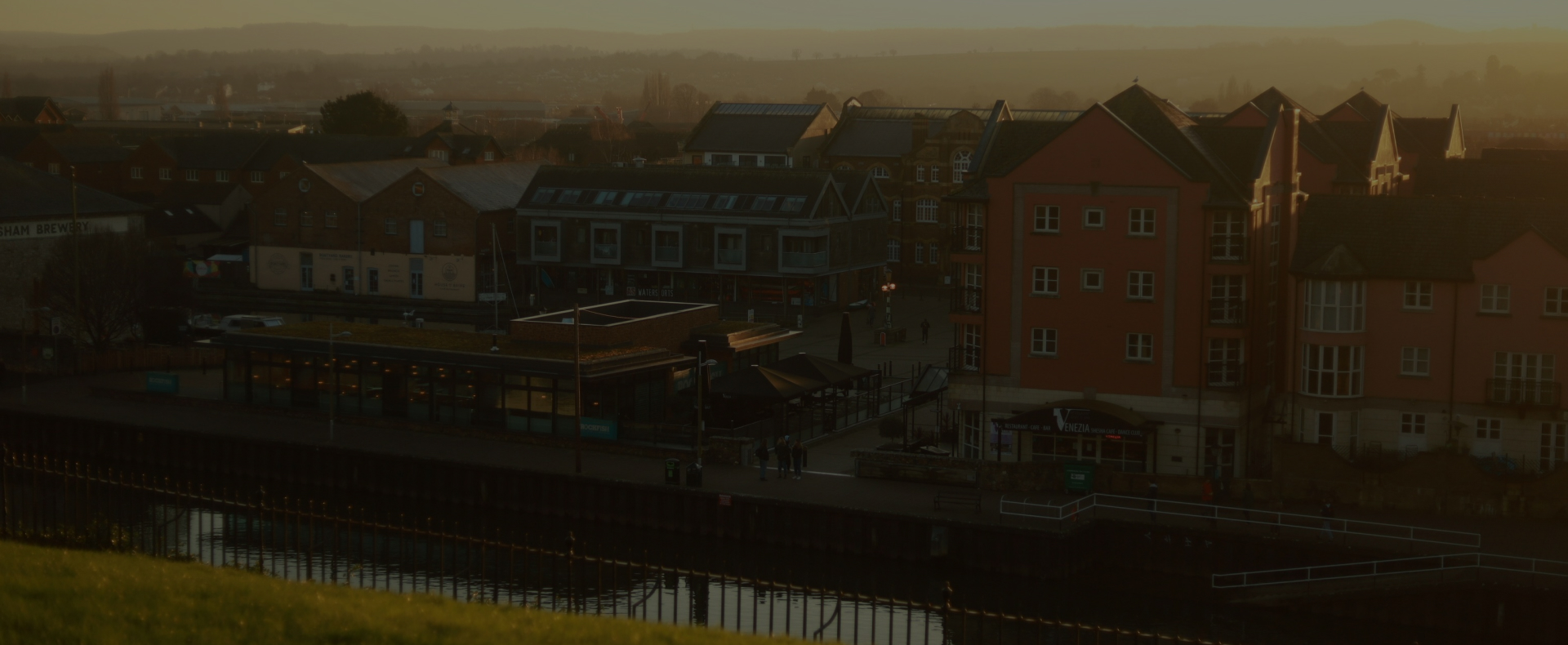 The quay in Exeter with railings and grass one side of the river and houses and other buildings on the other side.