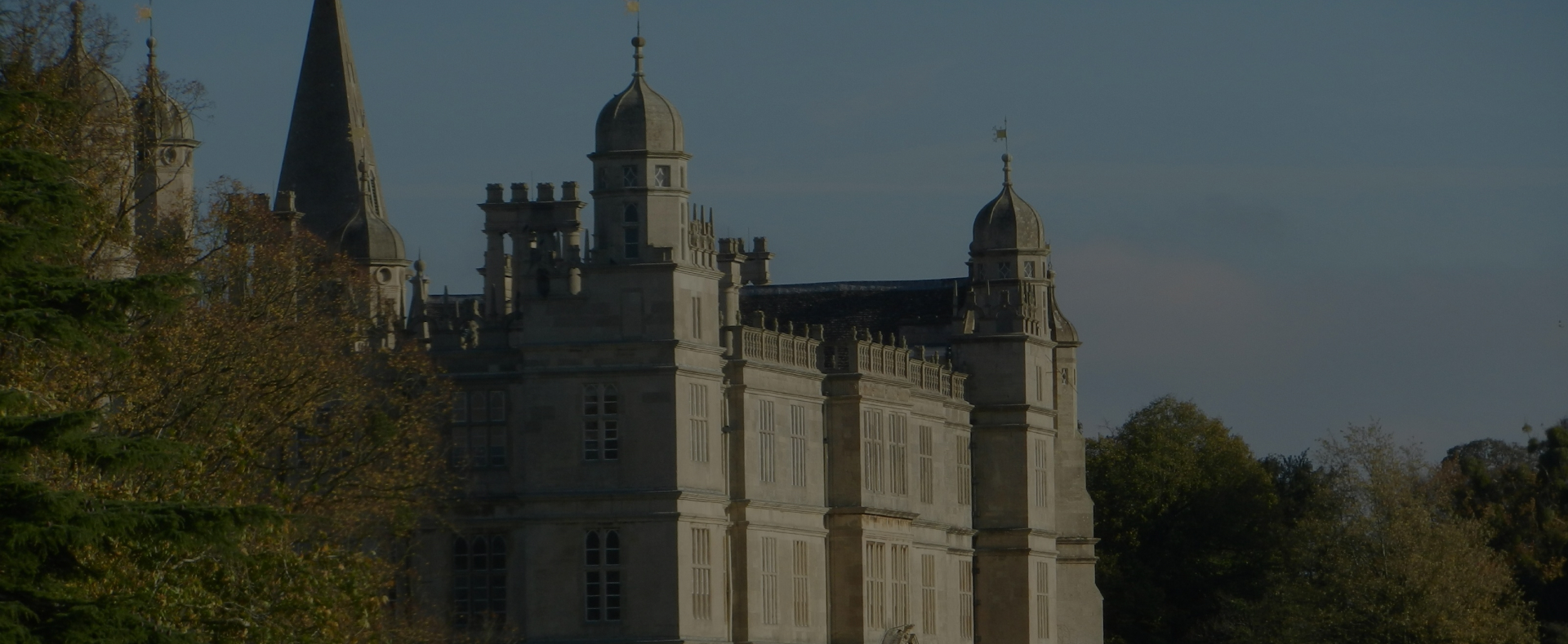 A large country house in Peterborough with spires and turrets with many windows surrounded by trees and a clear sky.
