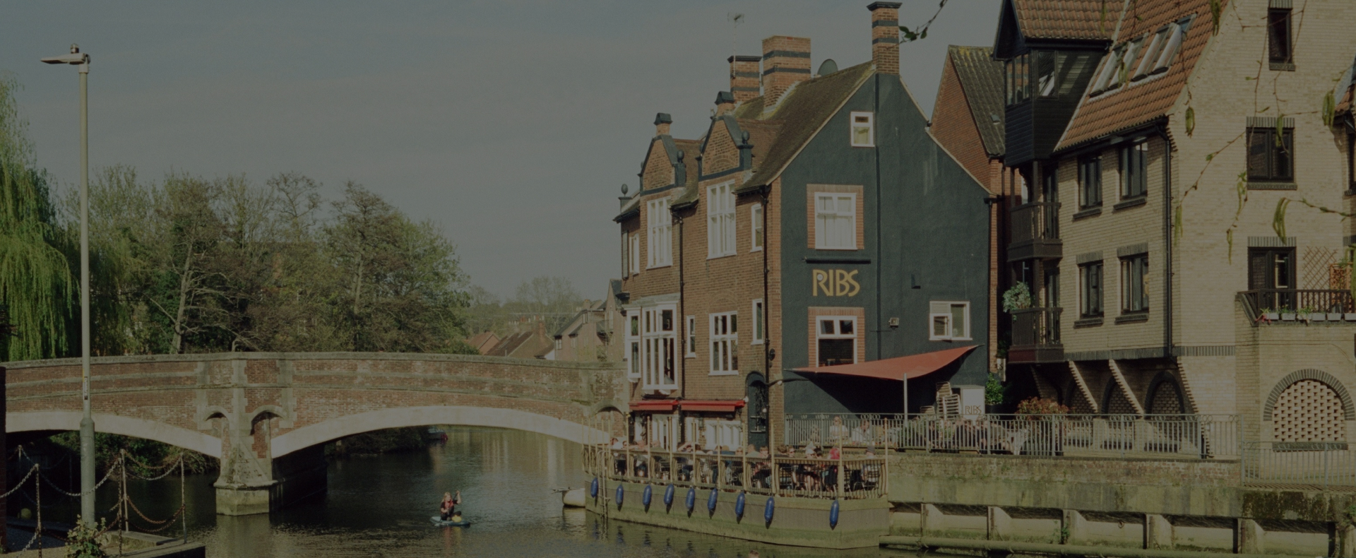 A bridge over the River Wensum in Norwich with a pub restaurant at one end surrounded by trees with a kayak in the water.